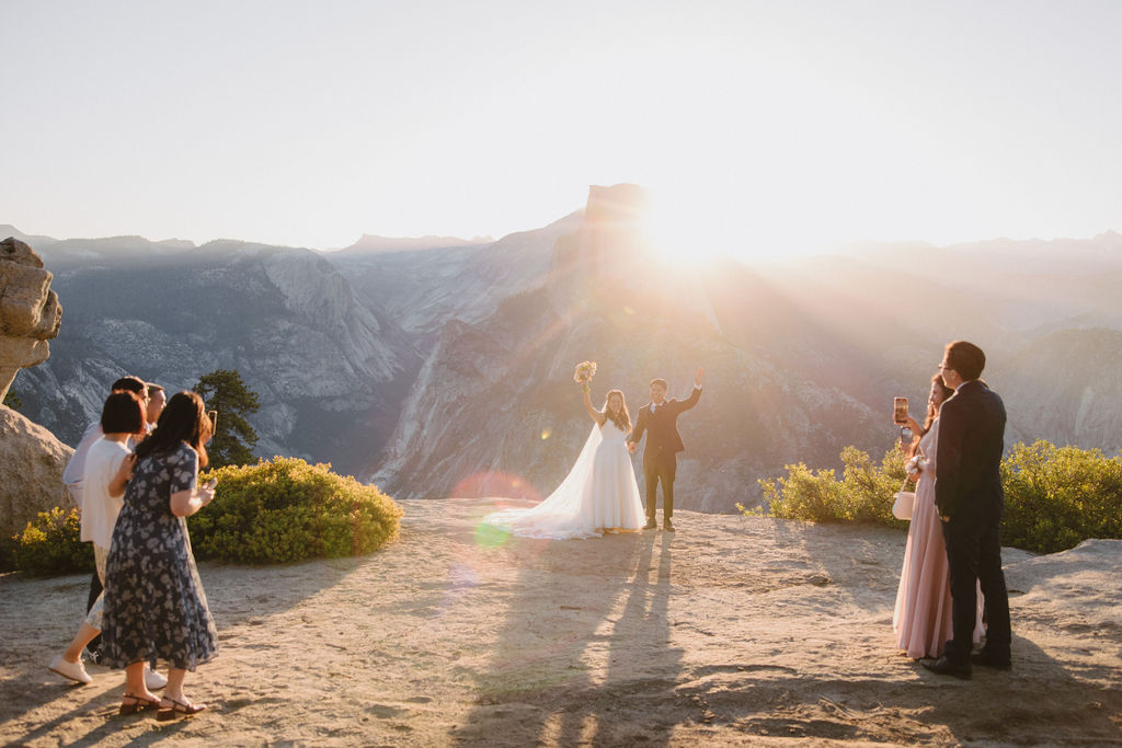 A wedding ceremony taking place on a cliffside with mountains in the background during sunset. The bride and groom stand at the center while a few guests are present, taking photos and watching.