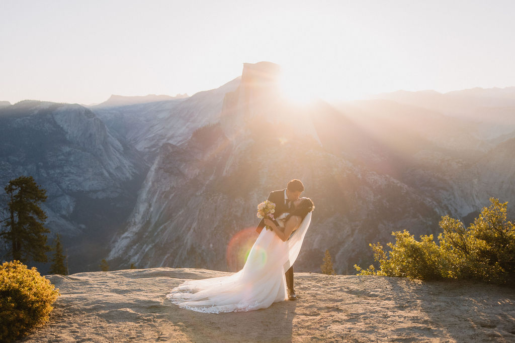 A couple in wedding attire embraces and kisses at sunset on a cliff with a mountain range in the background| tips for yosemite elopement 