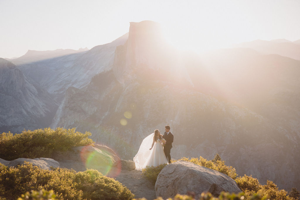 A bride and groom stand on a rocky outcrop, backlit by the setting sun, with mountain peaks and a hazy sky in the background in Yosemite national park