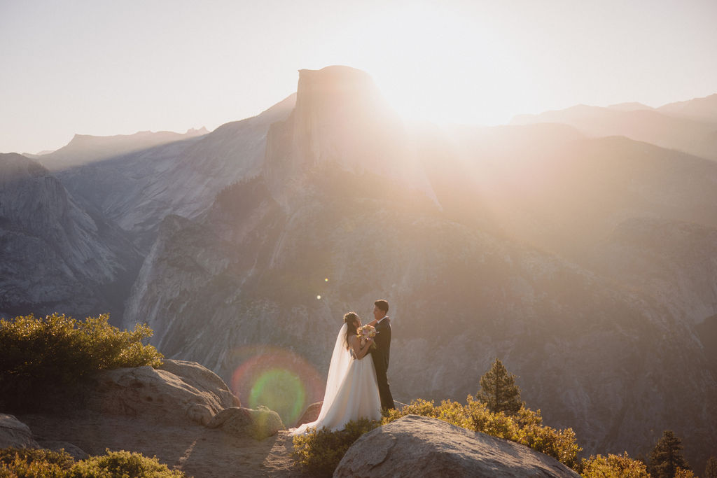 A bride and groom stand on a rocky outcrop, backlit by the setting sun, with mountain peaks and a hazy sky in the background in Yosemite national park