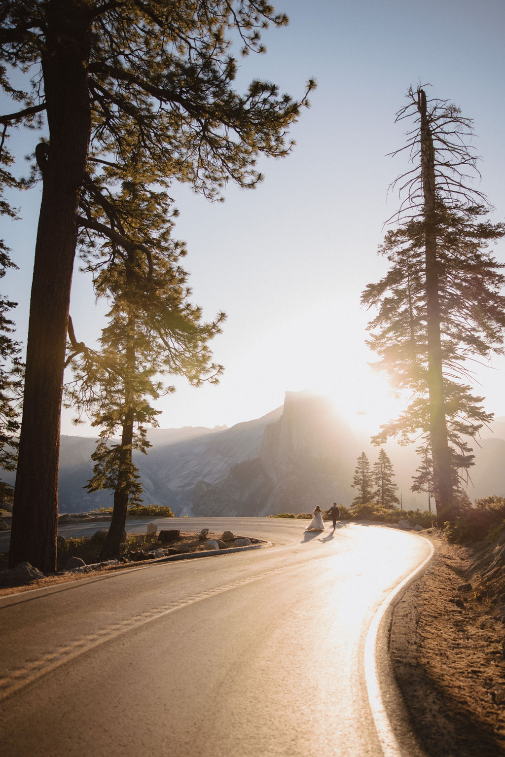 A couple in wedding attire holds hands while walking down a sunlit road surrounded by tall trees and mountains in the background at Yosemite