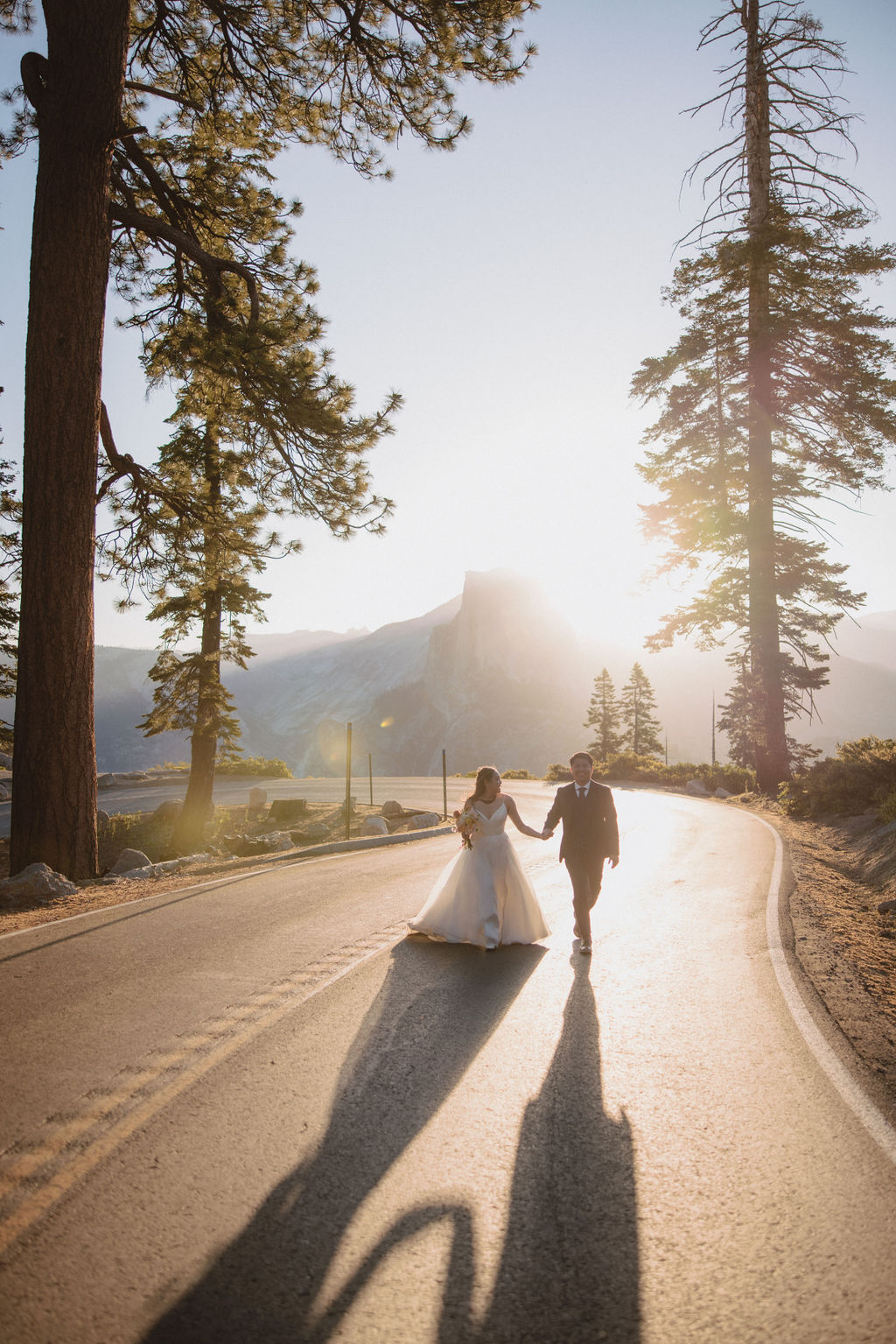 A couple in wedding attire holds hands while walking down a sunlit road surrounded by tall trees and mountains in the background at Yosemite