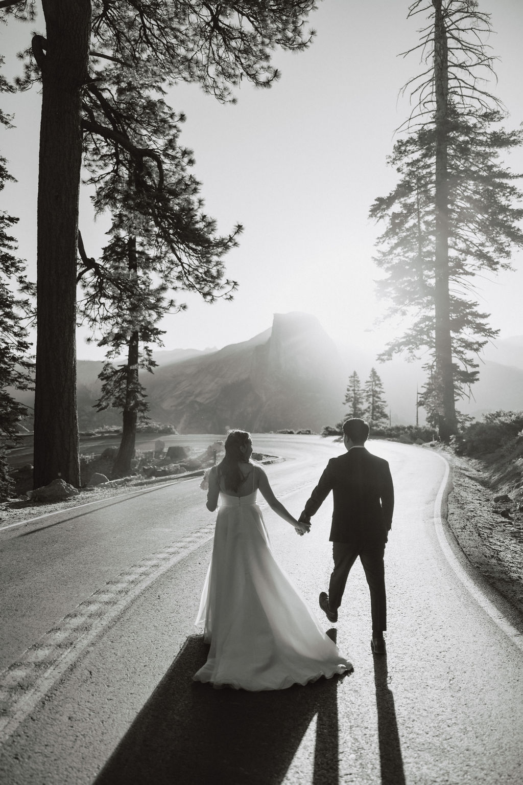 A couple in wedding attire holds hands while walking down a sunlit road surrounded by tall trees and mountains in the background at Yosemite