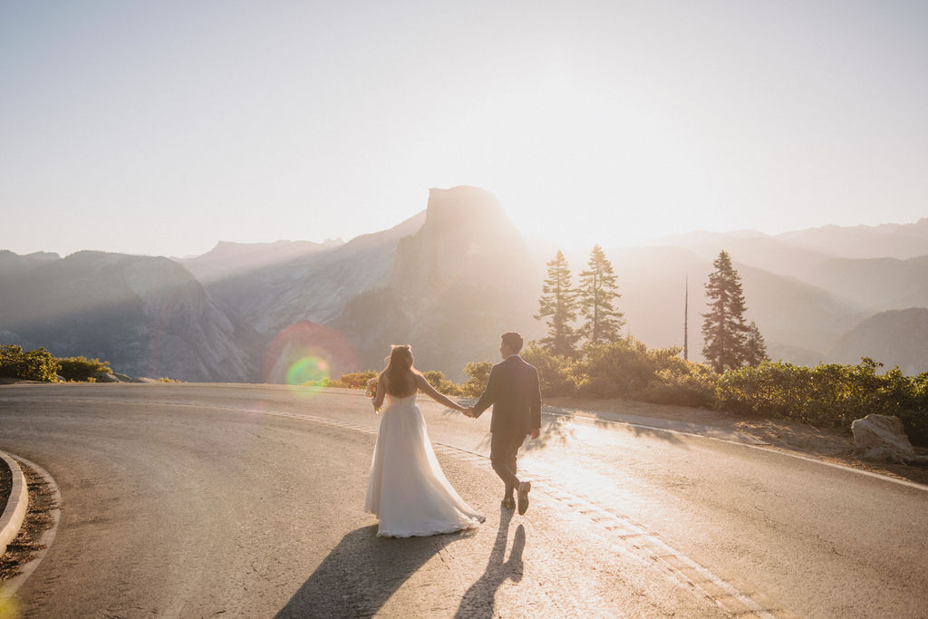 A couple in wedding attire holds hands while walking down a sunlit road surrounded by tall trees and mountains in the background at Yosemite