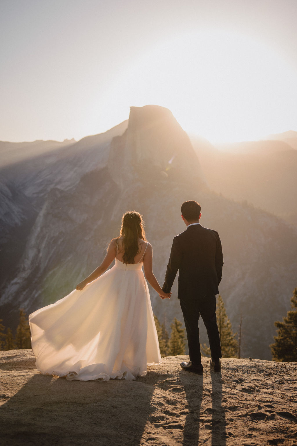 A couple in wedding attire stands hand-in-hand on a rocky cliff, facing a mountainous landscape at sunset for their elopement in Yosemite