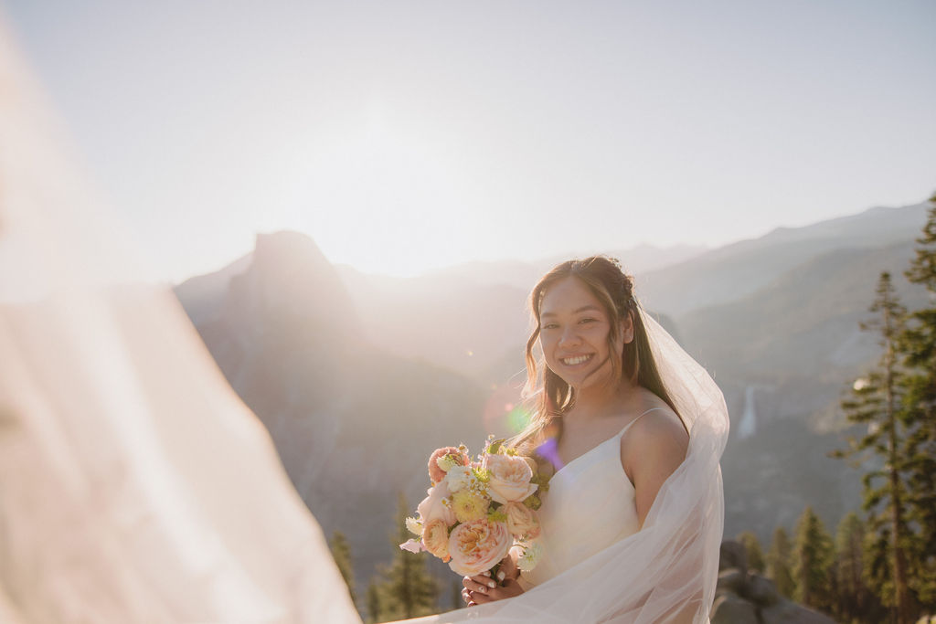 A bride in a white dress and veil smiles while holding a bouquet of flowers, standing outdoors with a mountainous landscape in the background.