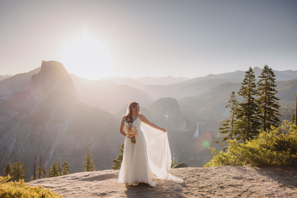 A bride in a white dress and veil smiles while holding a bouquet of flowers, standing outdoors with a mountainous landscape in the background.