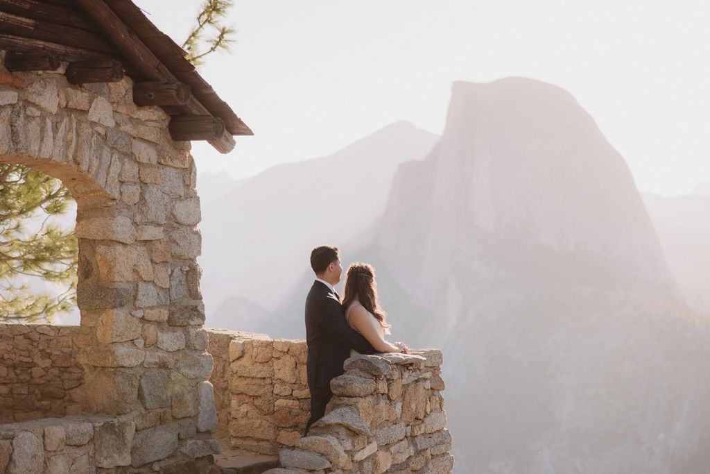 A couple stands on a stone balcony with a mountainous landscape in the background, gazing at the scenery in Yosemite