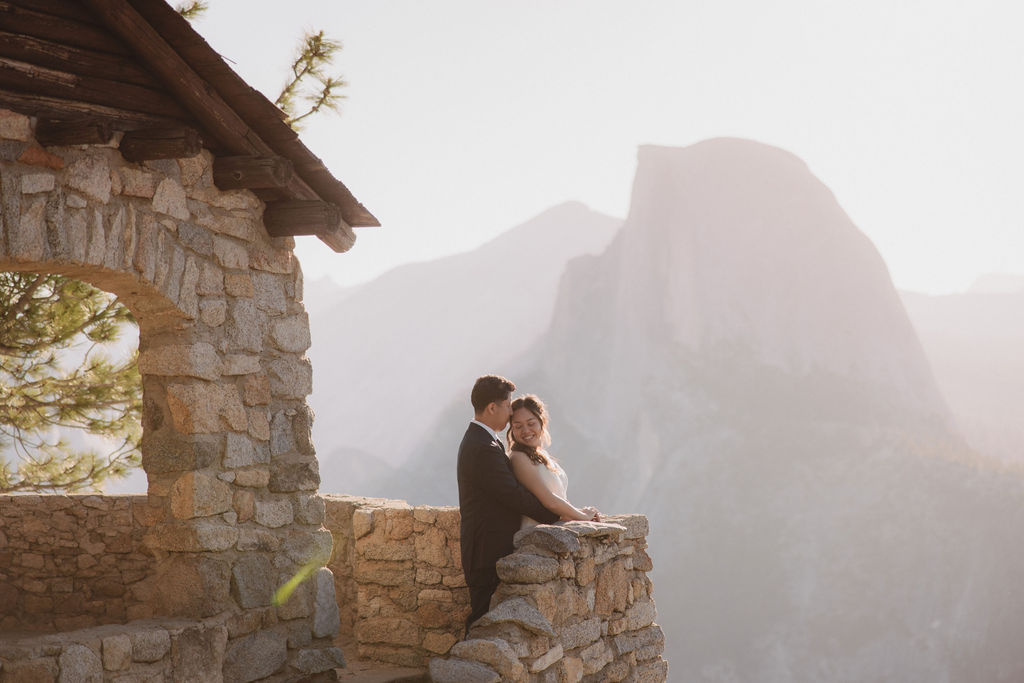 A couple stands on a stone balcony with a mountainous landscape in the background, gazing at the scenery in Yosemite