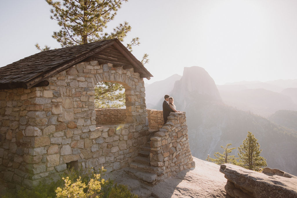 A couple stands on a stone balcony with a mountainous landscape in the background, gazing at the scenery in Yosemite