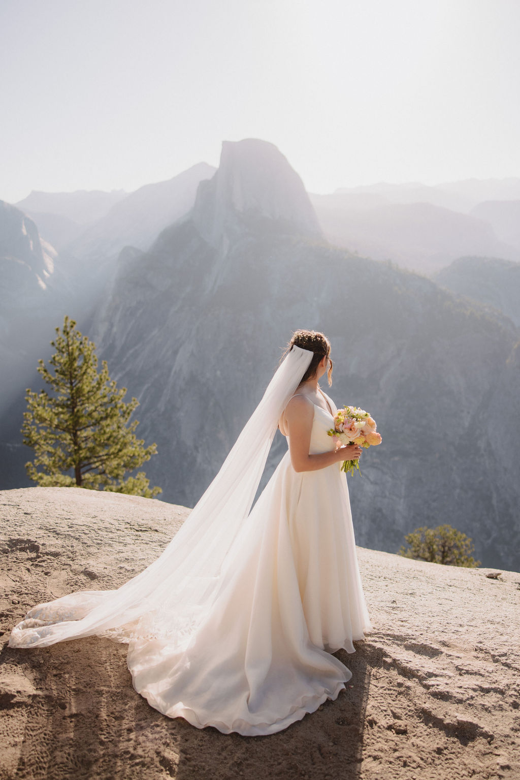 A bride in a white wedding dress and long veil stands on a mountain overlook holding a bouquet, with a backdrop of mountains and sunlight filtering through in Yosemite National Park