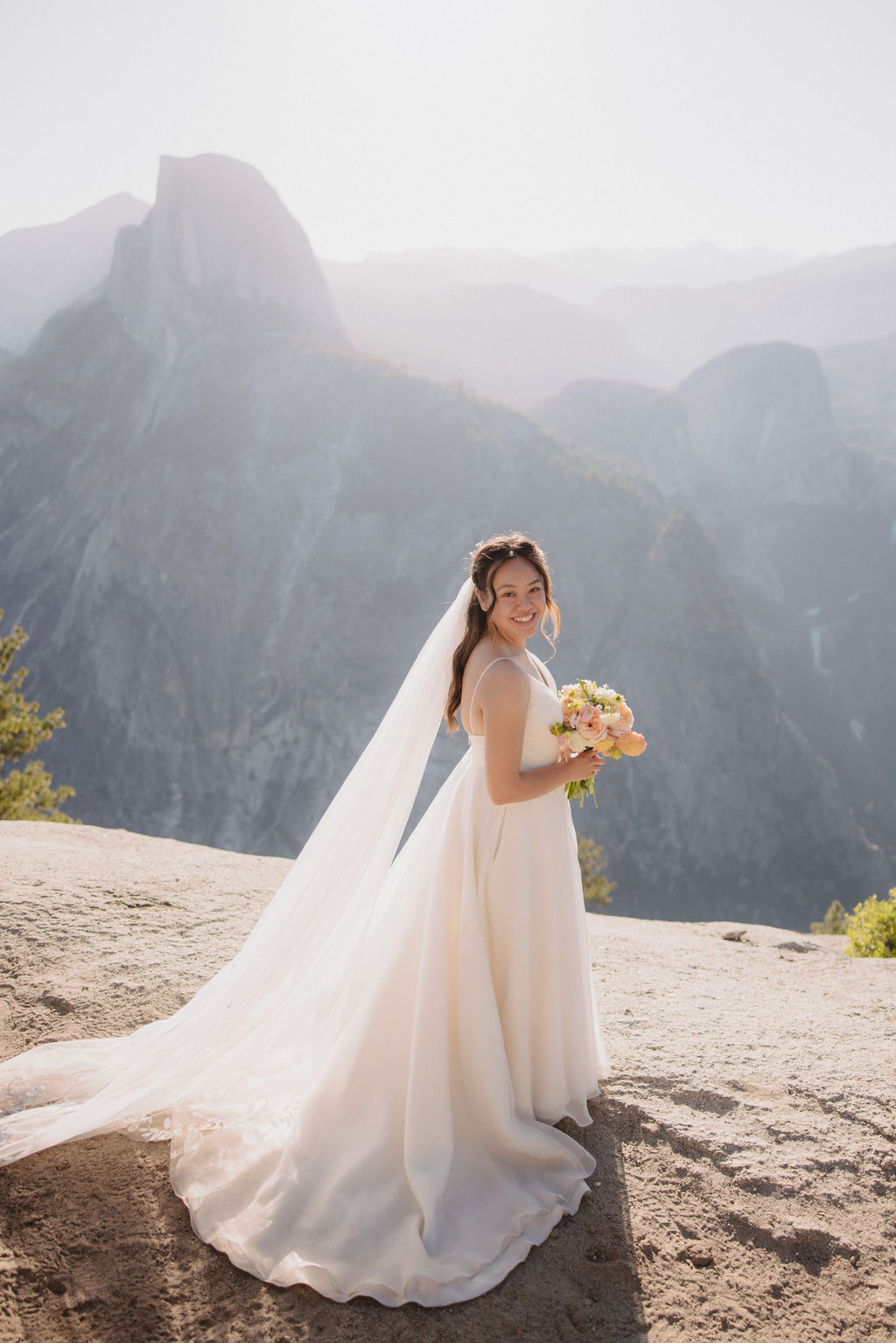 A bride in a white wedding dress and long veil stands on a mountain overlook holding a bouquet, with a backdrop of mountains and sunlight filtering through in Yosemite National Park