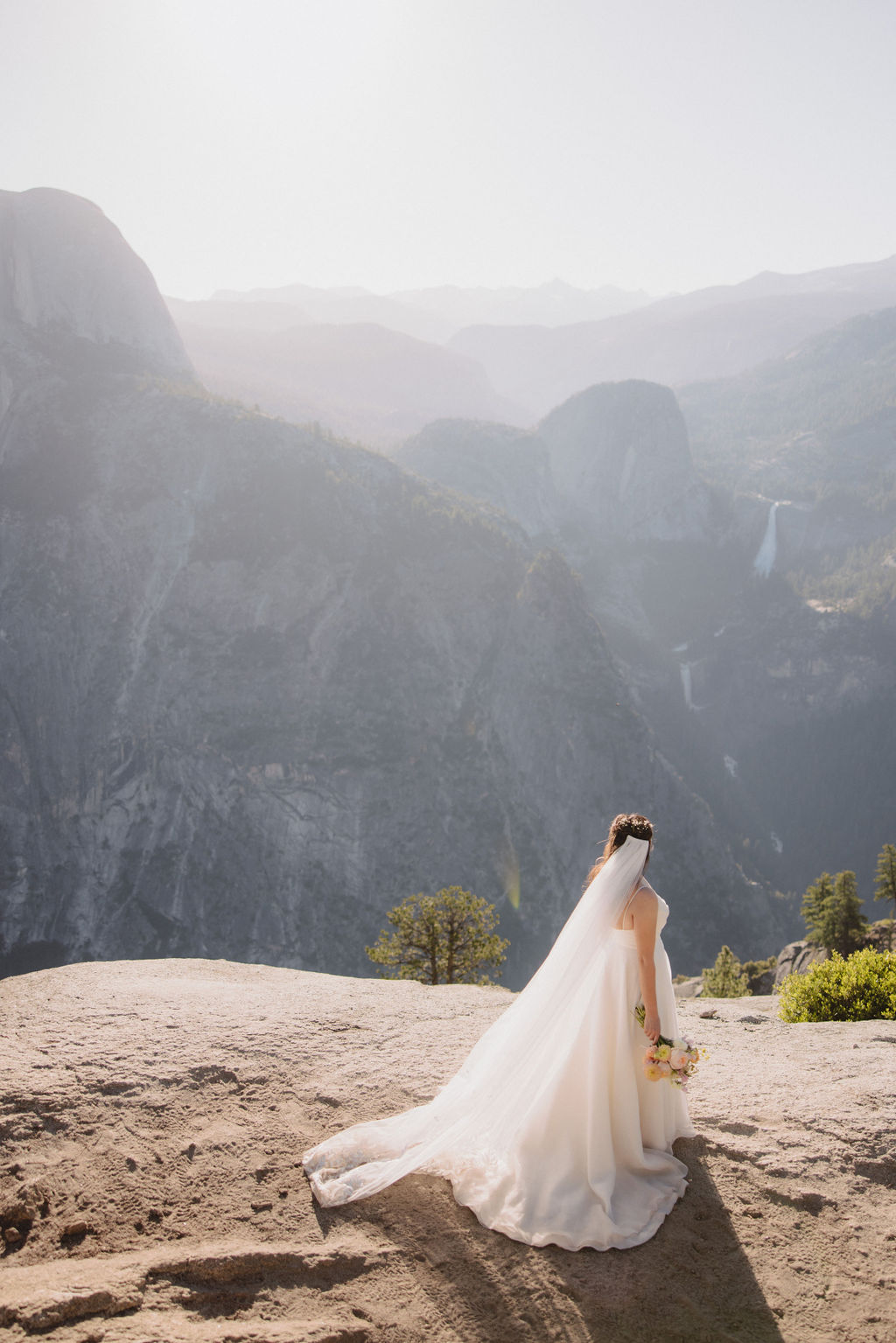 A bride in a white wedding dress and long veil stands on a mountain overlook holding a bouquet, with a backdrop of mountains and sunlight filtering through in Yosemite National Park