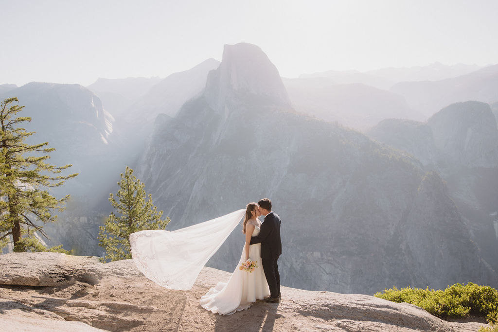 A bride in a white wedding dress and long veil stands on a mountain overlook holding a bouquet, with a backdrop of mountains and sunlight filtering through in Yosemite National Park