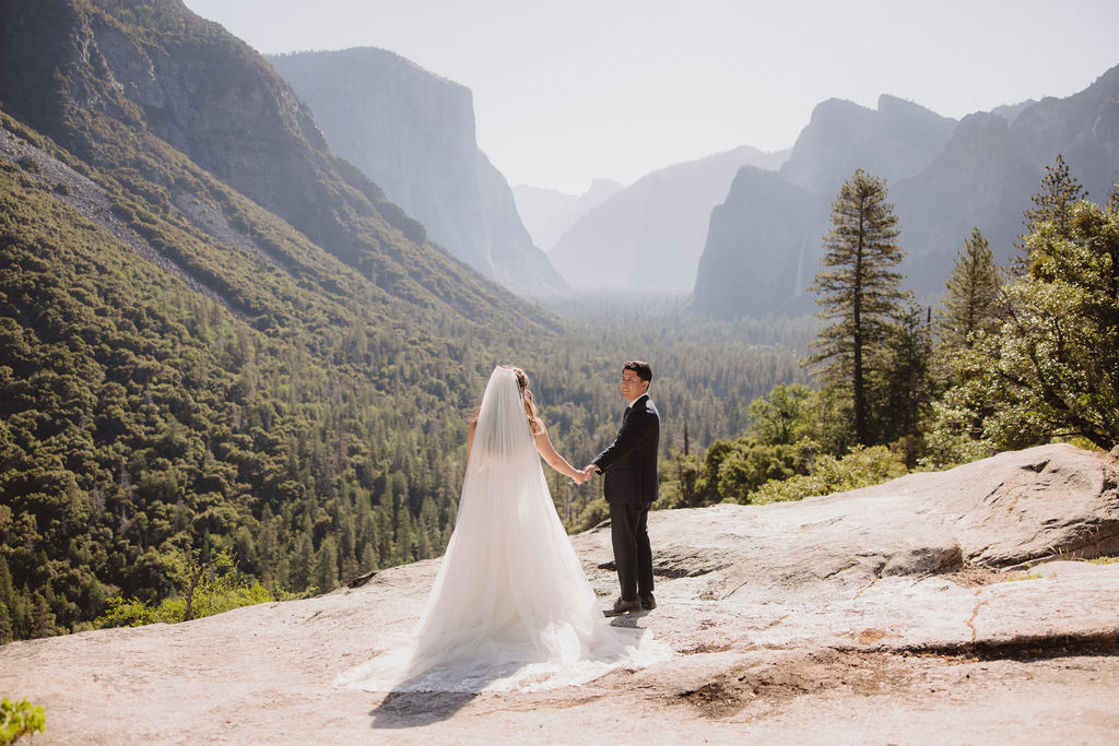 A bride and groom stand holding hands on a rocky outcrop with a scenic view of a valley surrounded by mountains and trees in Yosemite national park