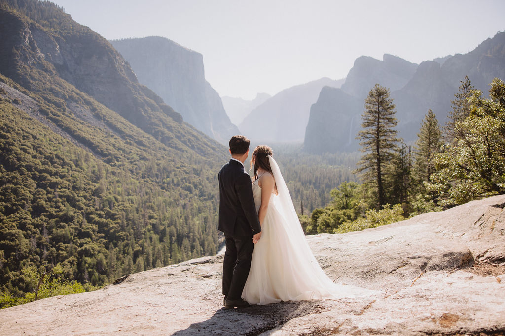 A bride and groom stand holding hands on a rocky outcrop with a scenic view of a valley surrounded by mountains and trees in Yosemite national park