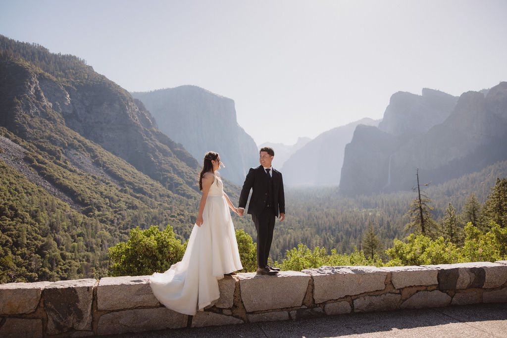 A bride and groom stand holding hands on a rocky outcrop with a scenic view of a valley surrounded by mountains and trees in Yosemite national park