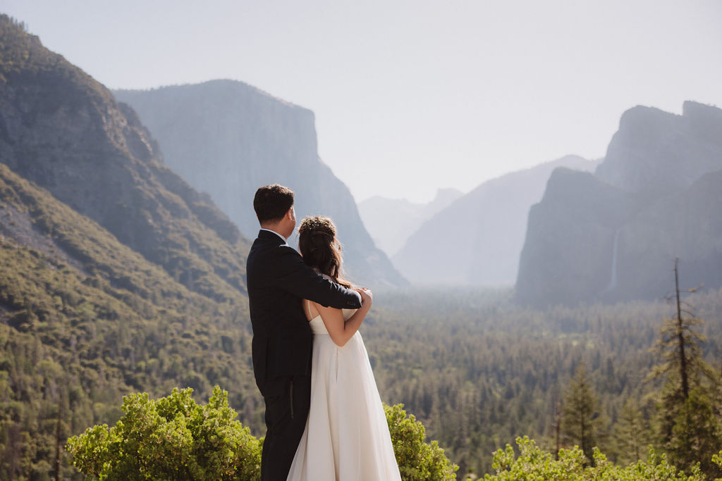 A bride and groom stand holding hands on a rocky outcrop with a scenic view of a valley surrounded by mountains and trees in Yosemite national park