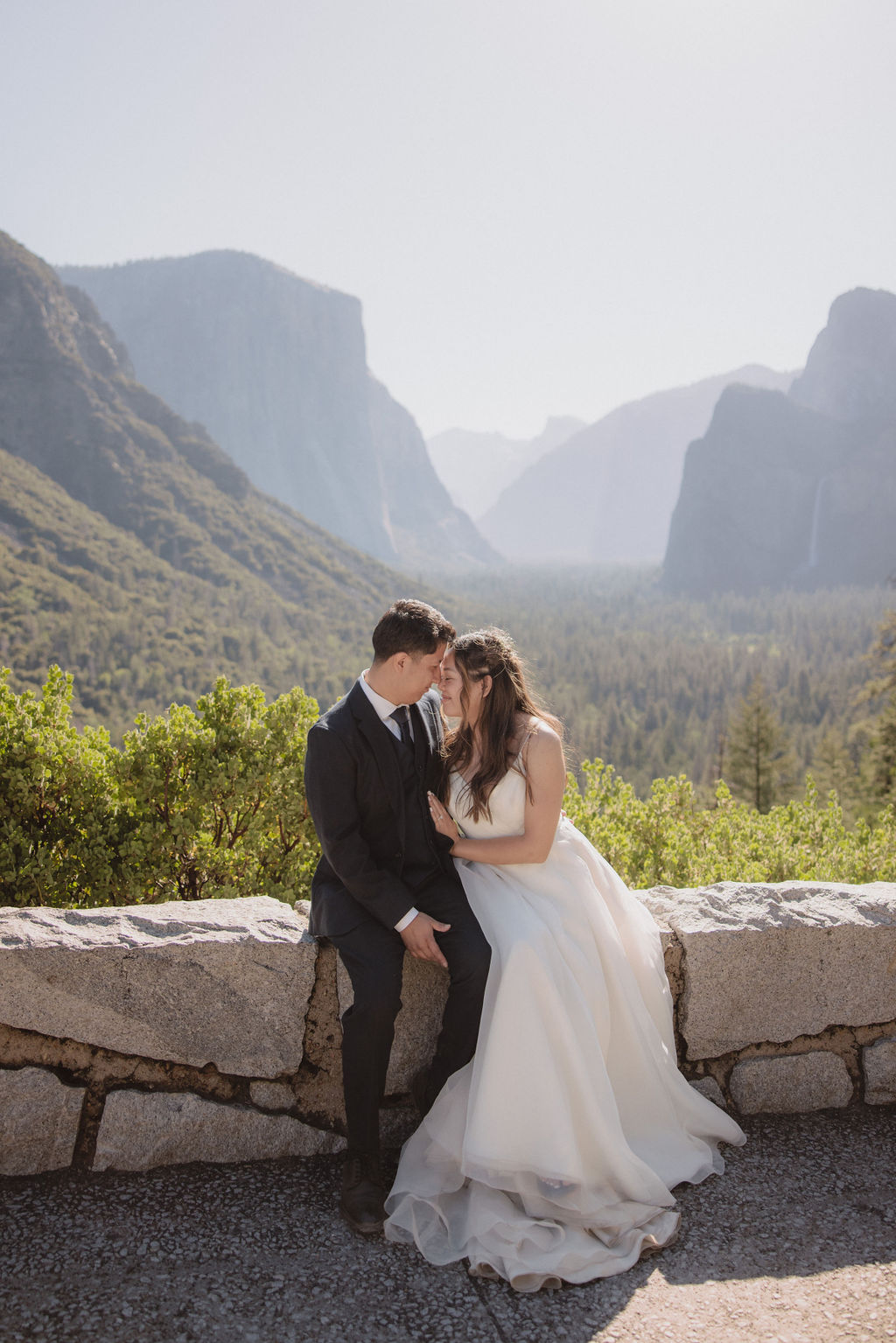 A bride and groom stand holding hands on a rocky outcrop with a scenic view of a valley surrounded by mountains and trees in Yosemite national park