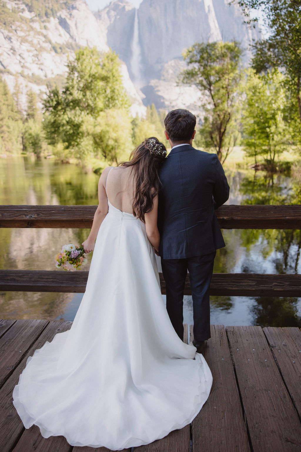 A couple in wedding attire holds hands and faces away, standing between two large trees in a forest meadow with mountains in the background.