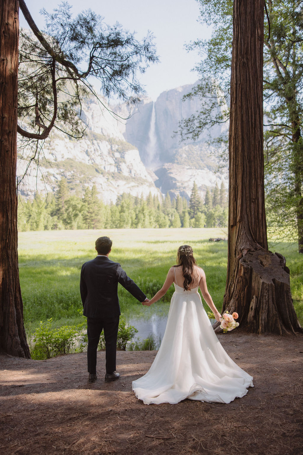 A couple in wedding attire holds hands and faces away, standing between two large trees in a forest meadow with mountains in the background.