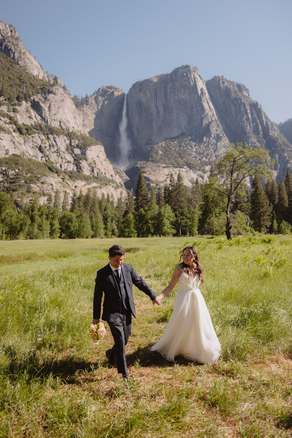 A bride and groom stand holding hands in a meadow with a mountain and waterfall in the background. The bride is holding a bouquet and both are looking away from the camera in Yosemite national park