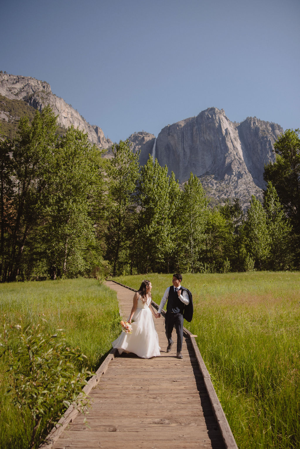 A bride and groom stand holding hands in a meadow with a mountain and waterfall in the background. The bride is holding a bouquet and both are looking away from the camera in Yosemite national park