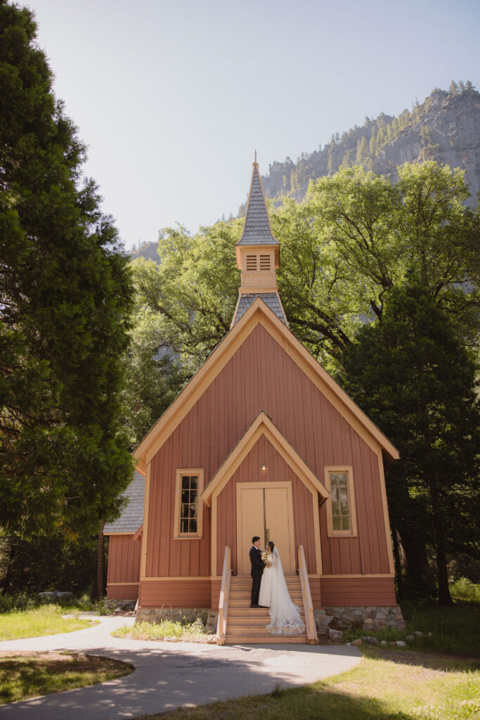 A couple in wedding attire stands at the entrance of a rustic, red-painted chapel surrounded by trees in Yosemite national park