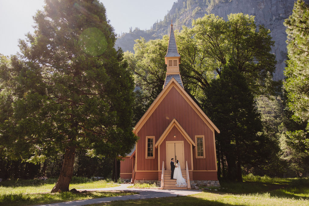 A couple dressed in wedding attire stands on the steps of a small, brown chapel surrounded by trees at Yosemite Chapel for their yosemite elopement 