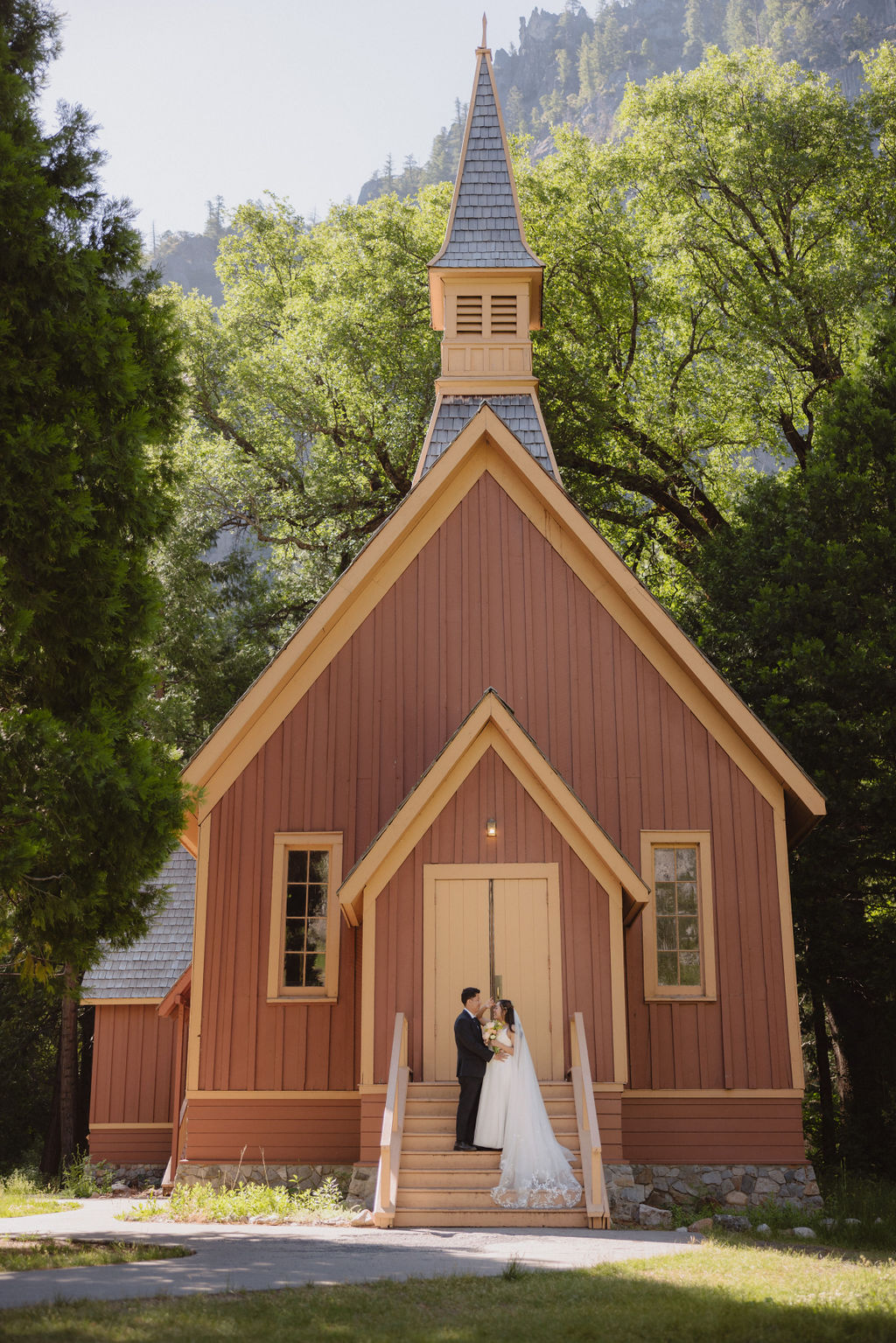 A couple in wedding attire stands at the entrance of a rustic, red-painted chapel surrounded by trees in Yosemite national park 