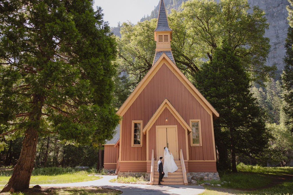 A couple in wedding attire stands at the entrance of Yosemite chapel surrounded by trees in Yosemite national park 