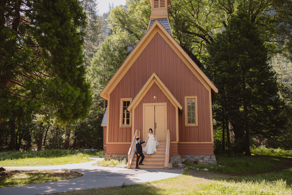 A couple in wedding attire stands at the entrance of Yosemite chapel surrounded by trees in Yosemite national park 