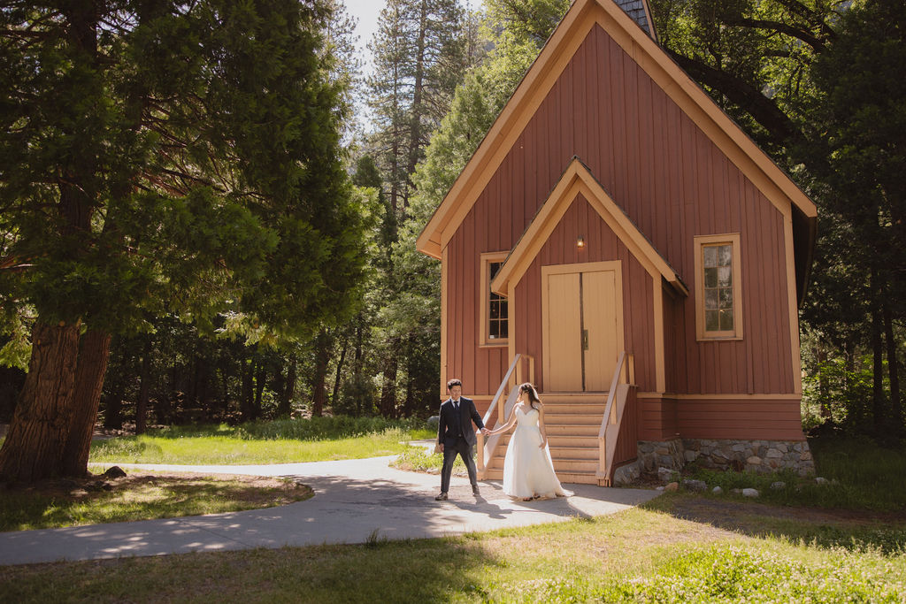 A couple in wedding attire stands at the entrance of Yosemite chapel surrounded by trees in Yosemite national park 