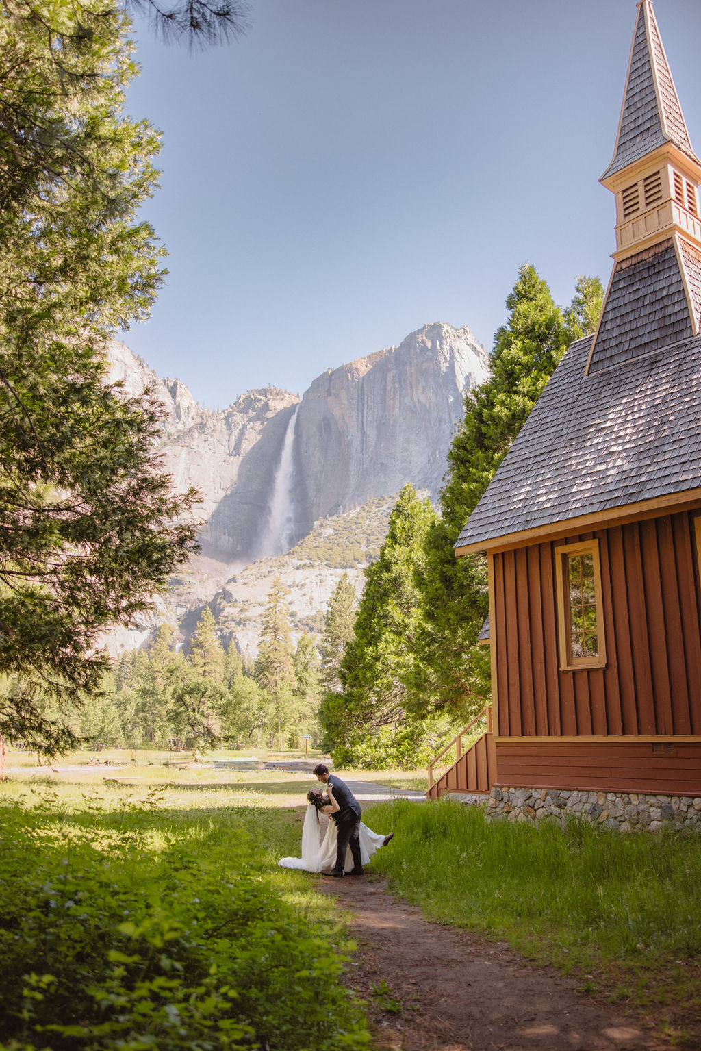 A couple in wedding attire stands at the entrance of Yosemite chapel surrounded by trees in Yosemite national park 