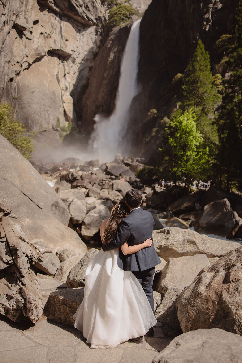 A couple in wedding attire stands embracing on rocky terrain with a waterfall cascading in the background at Yosemite falls 