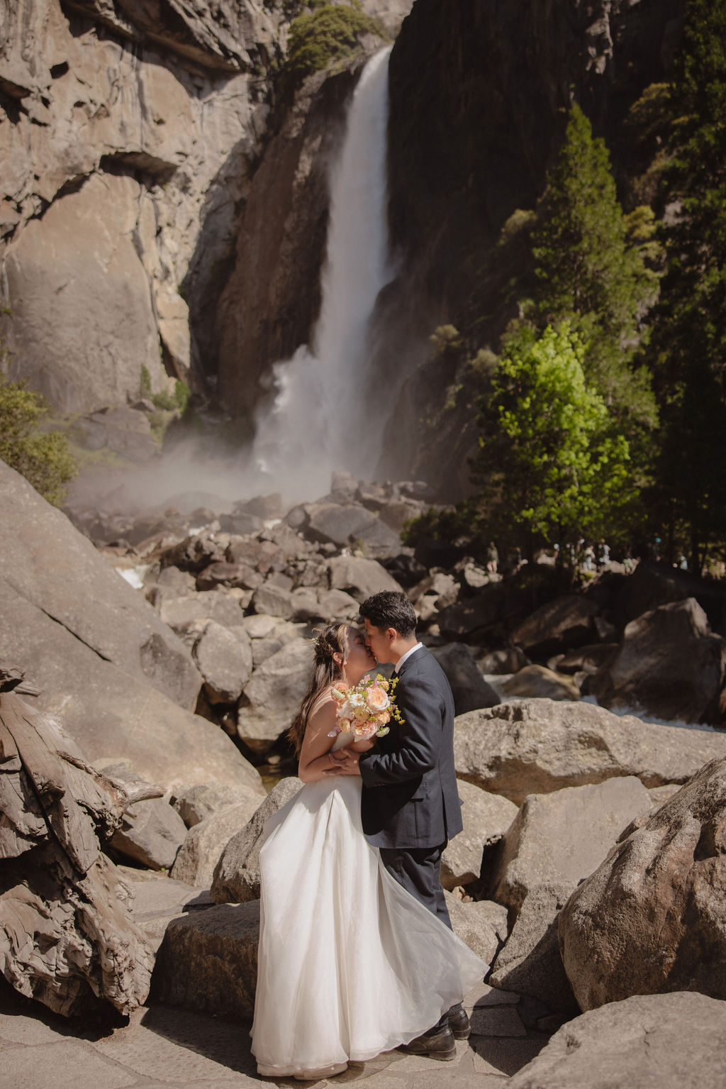 A couple in wedding attire stands embracing on rocky terrain with a waterfall cascading in the background at Yosemite falls 