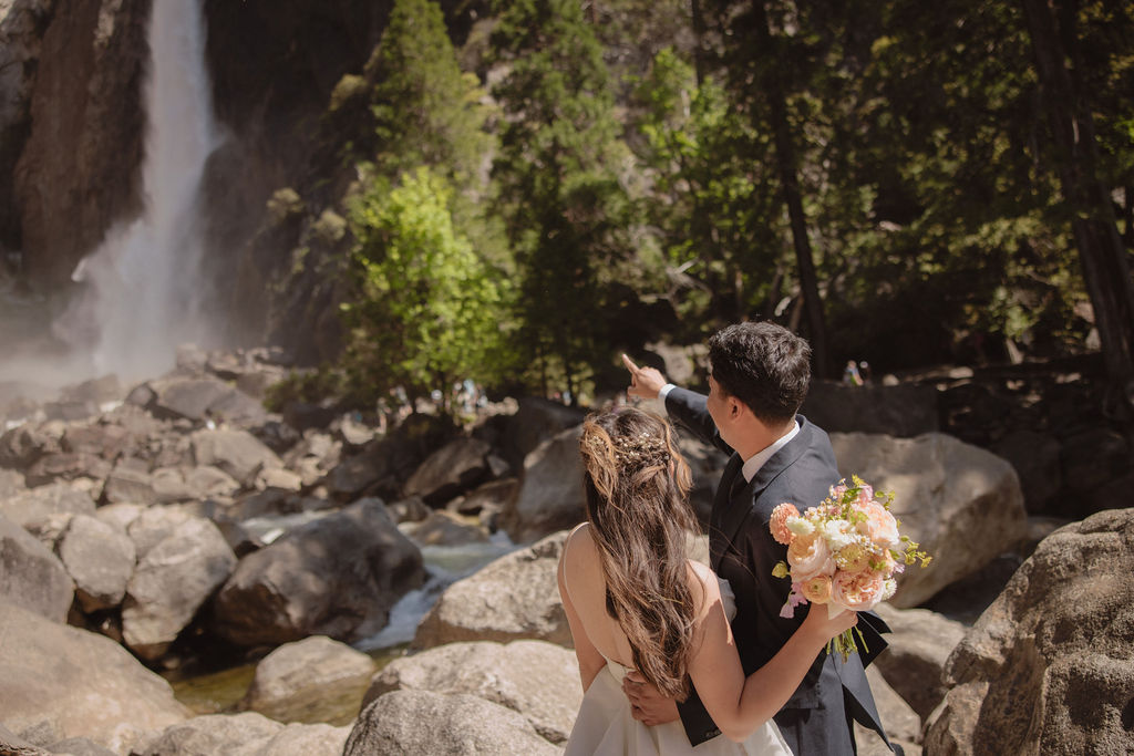 A couple in wedding attire stands embracing on rocky terrain with a waterfall cascading in the background at Yosemite falls 