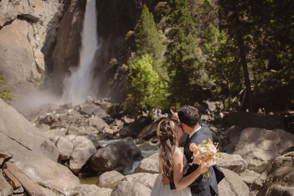 A couple in wedding attire stands embracing on rocky terrain with a waterfall cascading in the background at Yosemite falls 