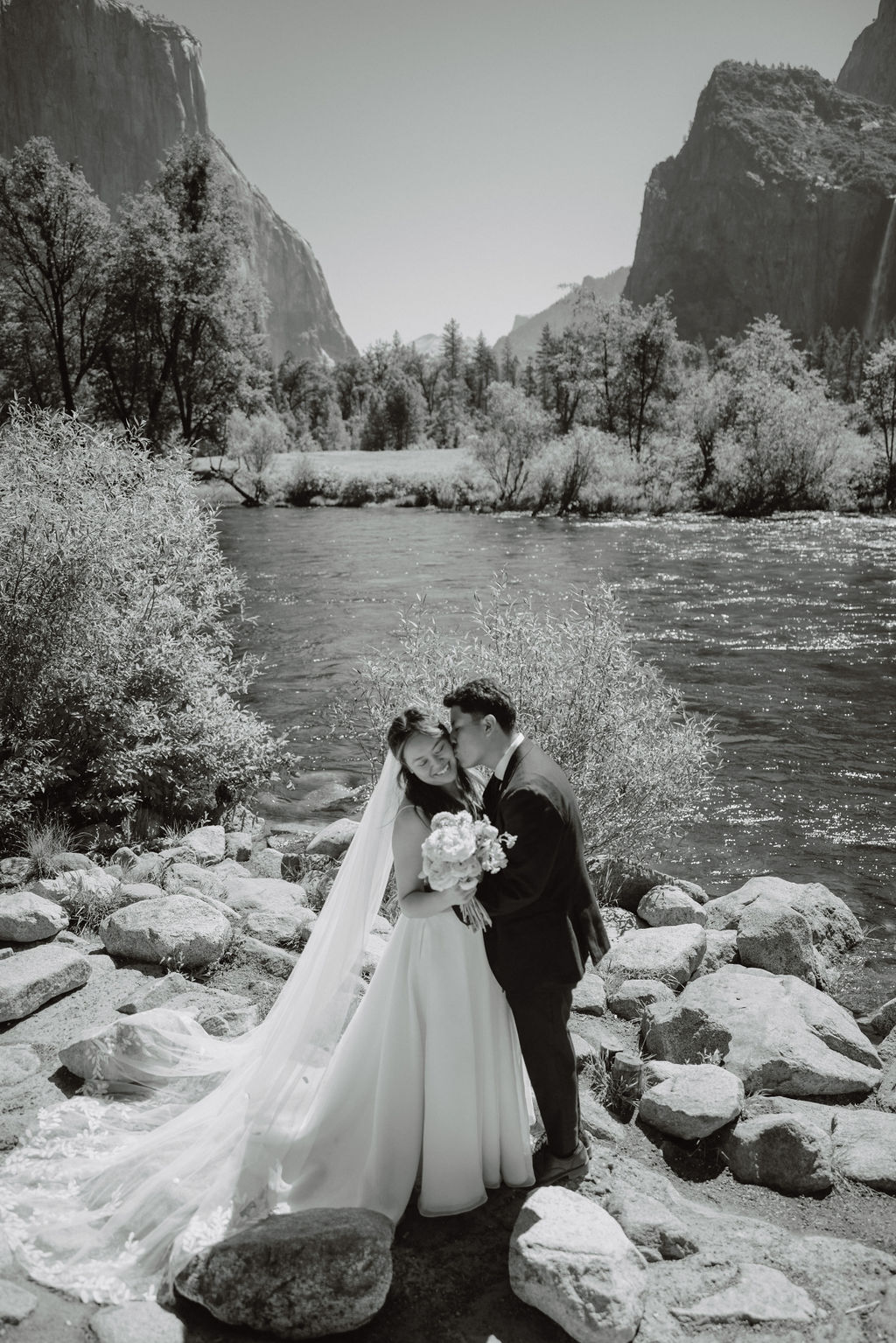 A couple in wedding attire stands embracing on rocky terrain with a waterfall cascading in the background at Yosemite falls 