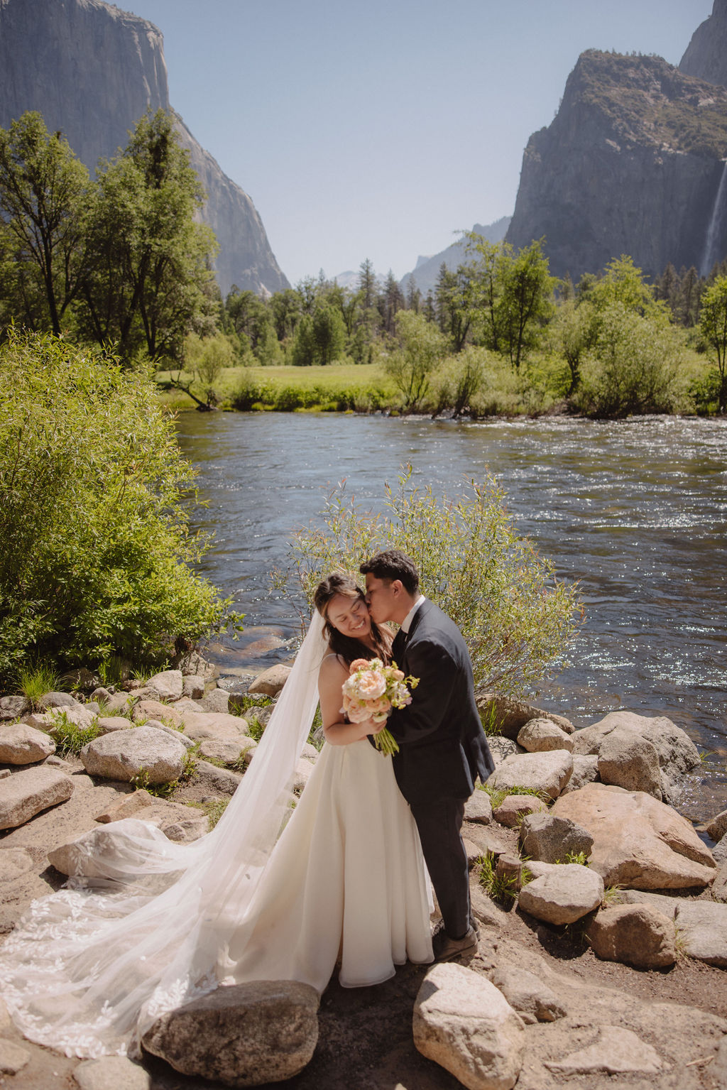 A couple in wedding attire shares a kiss beside a river with a scenic mountain and waterfall backdrop. The bride holds a bouquet.