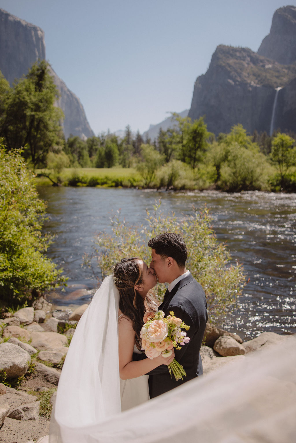 A couple in wedding attire shares a kiss beside a river with a scenic mountain and waterfall backdrop. The bride holds a bouquet.