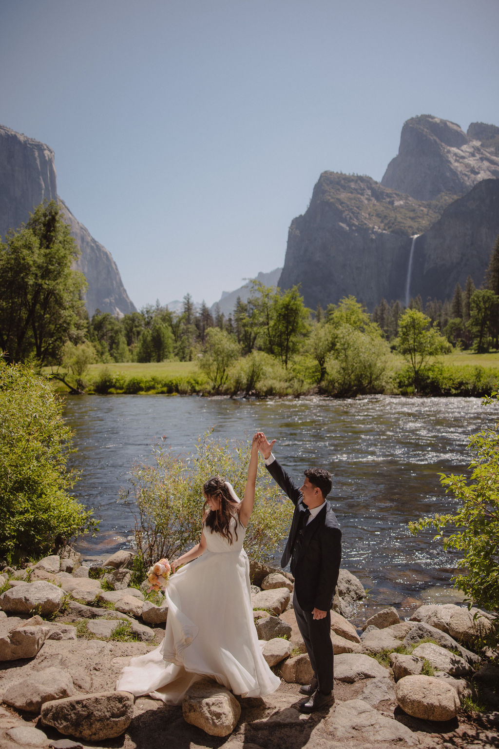 A couple in wedding attire shares a kiss beside a river with a scenic mountain and waterfall backdrop. The bride holds a bouquet.