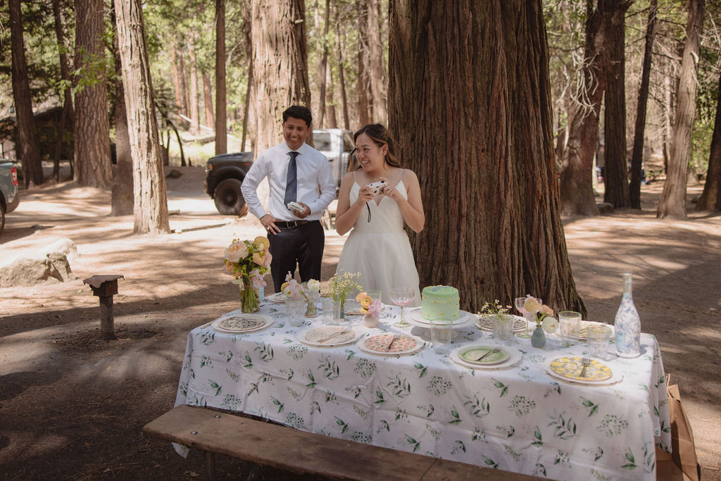 A woman in a white dress and a man in a white shirt and tie stand behind a table with various foods and cakes, set up outdoors among tall trees.