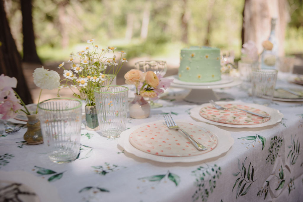 a table with various foods and cakes, set up outdoors among tall trees.