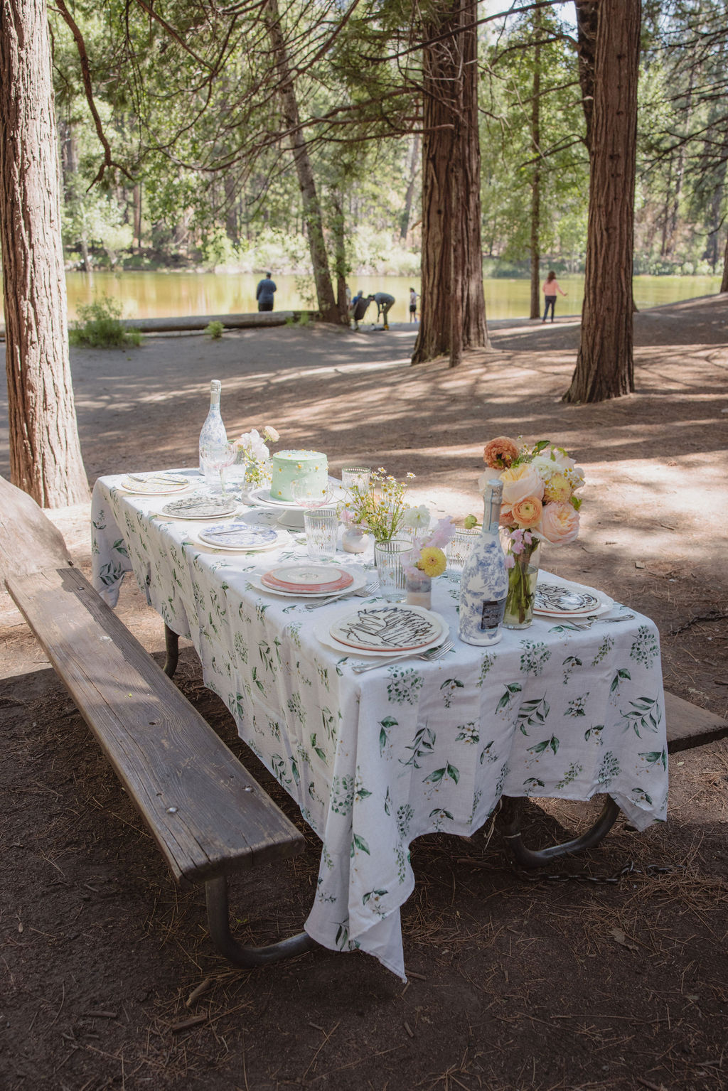 A picnic table set for an outdoor meal, covered with a white floral tablecloth and adorned with plates, glasses, silverware, and vases of flowers, amidst a wooded area near a body of water.