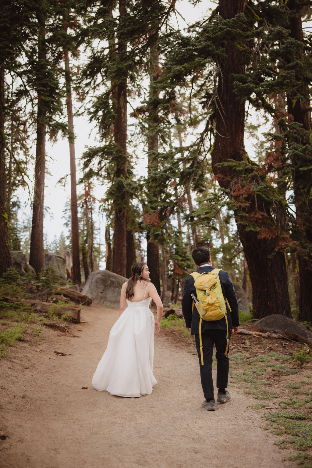 A person in a formal dress and another in casual attire with a backpack walk along a forest path surrounded by tall trees.