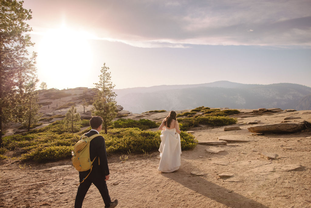 A couple in formal attire shares an intimate moment while standing on a log in a dense forest clearing with sunlight filtering through the trees in Yosemite