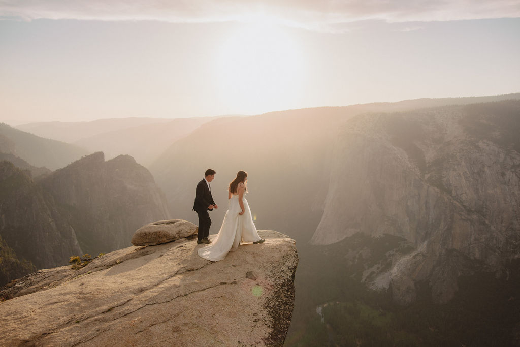 A couple in formal attire stands on the edge of a cliff, embracing and looking at each other, with vast mountainous scenery in the background under a hazy sky in Yosemite