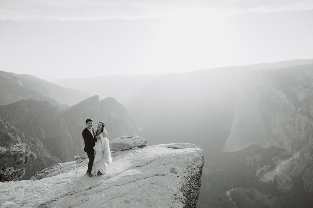 A couple in formal attire stands on the edge of a cliff, embracing and looking at each other, with vast mountainous scenery in the background under a hazy sky in Yosemite