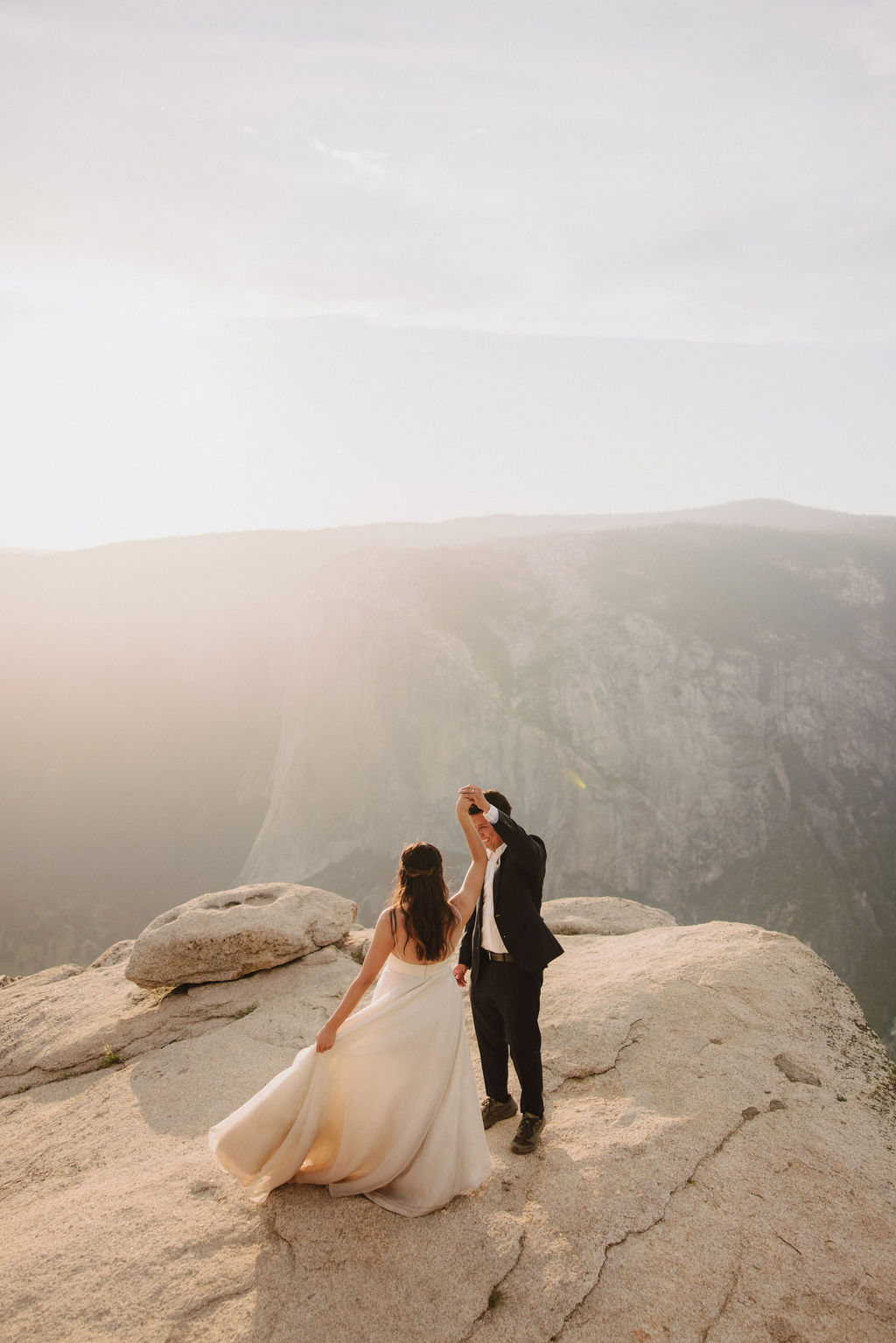 A couple in wedding attire stand on a rocky landscape. The woman in a white dress drinks from a bottle while the man in a black suit stands beside her holding a bottle.